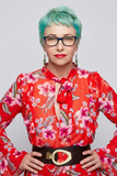 A woman with short turquoise hair, black glasses, and red lipstick wearing a red long-sleeve blouse with a pink and green floral print, a red bow tie at the collar, and a wide black belt with a gold buckle, posed against a light gray background.